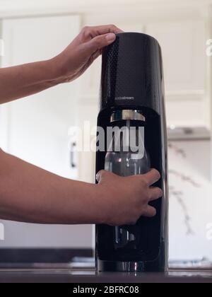 Latina woman holding a reusable Sodastream bottle as she carbonates the water it contains. Photographed at eye level in modern kitchen. Stock Photo