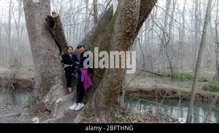 Two women in coats and sneakers posing by tree in the forest by trail n Orange County, North Carolina Stock Photo