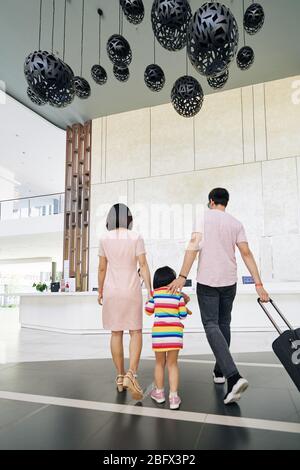 Family of three with big suitcase walking to hotel reception, view from the back Stock Photo