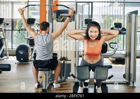 Cheerful young pretty Asian woman doing hypertension in gym when her boyfriend doing exercise at lat pulldown machine Stock Photo