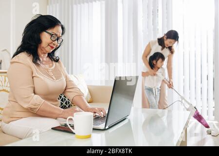 Smiling senior Asian woman working on laptop at home, her daughter and granddaughter vacuum cleaning floor in background Stock Photo