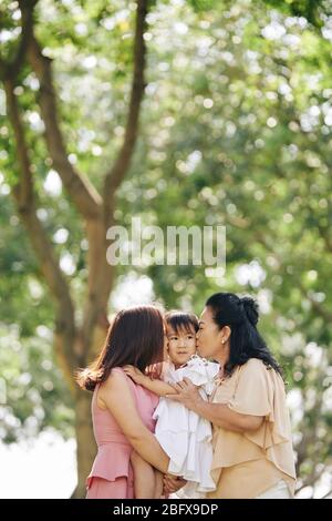 Beautiful granny and mother hugging and kissing little girl when standing in park Stock Photo