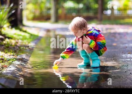 Kid playing in the rain in autumn park. Child jumping in muddy puddle on rainy fall day. Little boy in rain boots and red jacket outdoors in heavy sho Stock Photo