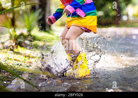 Kid playing in the rain in autumn park. Child jumping in muddy puddle on rainy fall day. Little girl in rain boots and rainbow jacket outdoors in heav Stock Photo