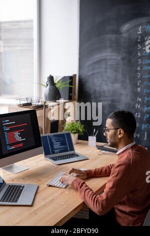 Concentrated young Arabian programmer in eyeglasses sitting at table and typing on keyboard among three computer while coding script Stock Photo