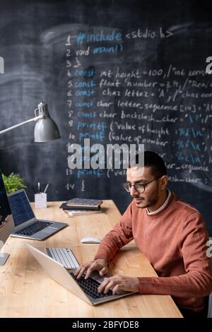 Talented young middle-eastern coder with beard sitting at desk and using laptop while working in office with blackboard Stock Photo