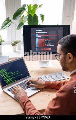 Over shoulder view of young web developer sitting at desk and working with laptop while creating computer algorithm Stock Photo