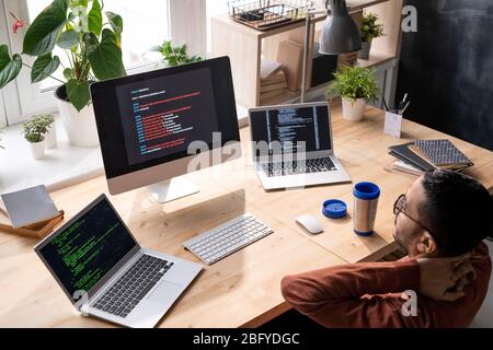 Above view of puzzled middle-eastern programmer in glasses sitting in front of computers and analyzing web script Stock Photo