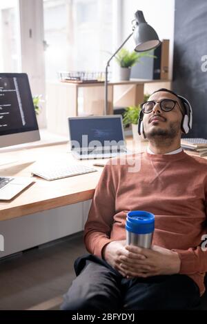 Exhausted young Arabian coder in headphones and glasses sleeping with thermos cup in hands at workplace Stock Photo