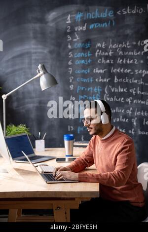 Young Arabian programmer focused on web code sitting at wooden table and typing on laptop while listening to audio in headphones Stock Photo