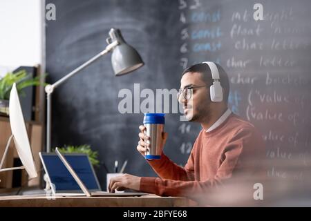 Serious young Arabian programming specialist in wireless headphones drinking hot tea from thermos cup and using laptop in office Stock Photo