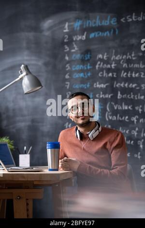 Portrait of smiling handsome young middle-eastern web developer in glasses sitting at table with laptop and thermos cup in office Stock Photo