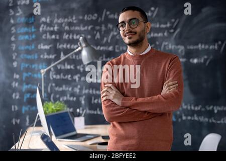 Pensive confident young middle-eastern programmer with beard standing with crossed arms in office with computer language code on blackboard Stock Photo