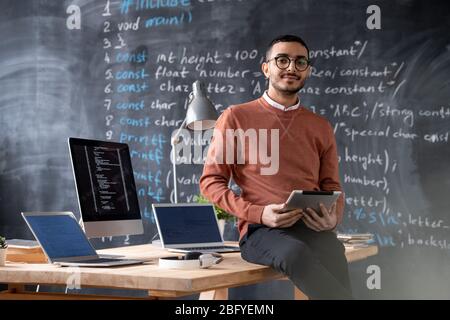Portrait of confident young Arabian coder in glasses sitting on table and using tablet against blackboard with computer script Stock Photo