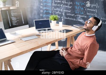 Tired young middle-eastern coder in headphones sitting in relaxed pose on chair while taking break at workplace Stock Photo