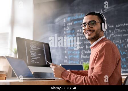 Portrait of smiling young coding specialist in wireless headphones sitting at desk with modern computers and checking smartphone in office Stock Photo