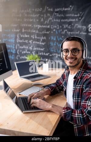 Portrait of smiling young network system administrator in wireless headphones sitting at desk and typing on laptop in office Stock Photo