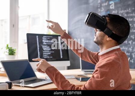 Concentrated young Arabian man sitting in computer office and developing app in VR goggles Stock Photo