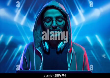 Young hacker in hoody sweatshirt and glasses breaking into computer system against blue illuminated background with geometric lines Stock Photo