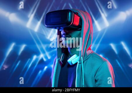 Hipster young man with stubble immersed in virtual reality wearing hoody sweatshirt watching 3D movie against blue luminous background Stock Photo