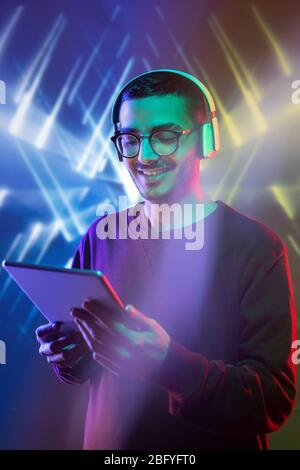Excited young Arabian man addicted to gadget listening to music in headphones playing online game against bright geometric background Stock Photo