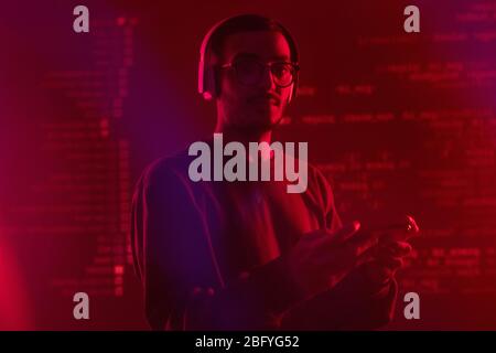 Portrait of hipster Arabian man in wireless headphones coding app script on smartphone in red light, coding background Stock Photo