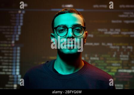Portrait of serious young Arabian programmer in eyeglasses standing against coding background Stock Photo