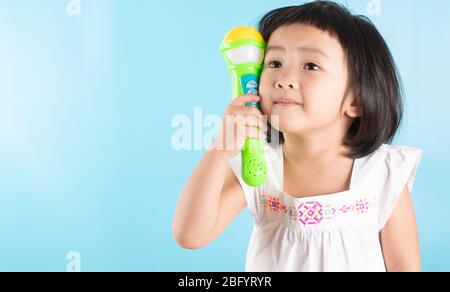 A cute Asian girl is holding a plastic microphone and singing with fun. On a blue background and copy space. Stock Photo