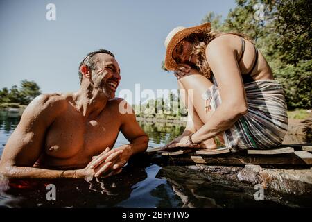 Mature man in lake with woman sitting at the pier. Couple enjoying on their camping vacation at the lake. Stock Photo