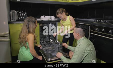 Mother and father teaching daughter learning to use dishwasher. Mistress girl children loading putting dirty dishes in automatic dishwasher. Stock Photo