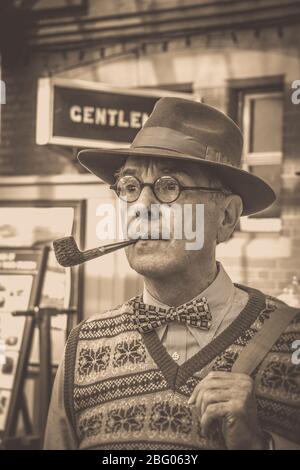 Old-fashioned vintage sepia close up of isolated man with pipe & trilby hat vintage train station, Severn Valley Railway 1940s wartime summer event UK. Stock Photo