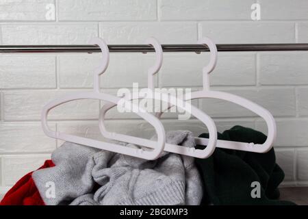 Empty white hangers, and under them a pile of clothes. Fashion, style. Sweaters in red, green and melange colors. White brick wall. Mess, home routine Stock Photo