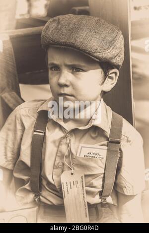 Old-fashioned, sepia portrait of evacuee boy in flat cap & trouser braces looking unhappy at vintage railway station, 1940s ww2 summer event, UK. Stock Photo