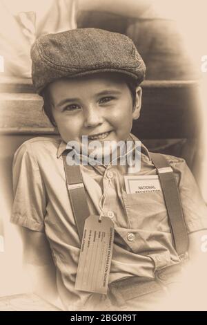 Old-fashioned, vintage sepia close-up portrait of smiling evacuee boy in flat cap & braces isolated at vintage railway station, 1940s summer event, UK. Stock Photo