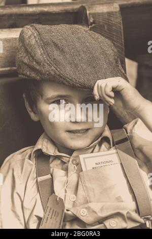 Old fashioned sepia close up of evacuee boy, vintage child in flat cap & braces, isolated at vintage railway station, 1940s ww2 summer event, UK. Stock Photo