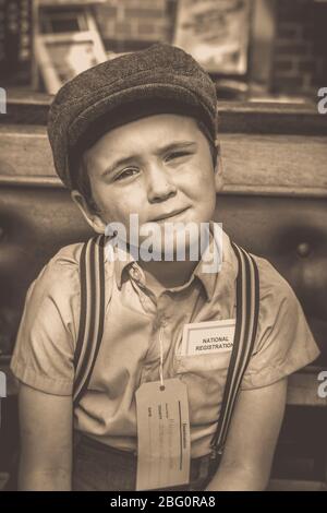 Old-fashioned, vintage sepia close up portrait of evacuee boy in flat cap & braces looking anxious at vintage railway station, 1940s summer event, UK. Stock Photo