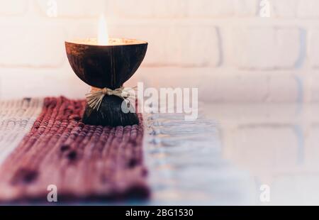 Burning spa aroma candle in coconut shell on a knitted multicolored rug opposite the white wall, cozy home interior Stock Photo