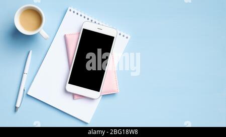 Modern office desk with smartphone with blank mockup screen, cup of coffee, blank paper notepad, document on blue background. Top view with copy space Stock Photo