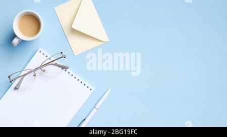 Modern workspace with cup of coffee, blank notepad, envelope, glasses on blue background. Minimal style feminine office desk, business and education c Stock Photo