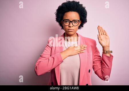 Young beautiful African American afro businesswoman with curly hair wearing pink jacket Swearing with hand on chest and open palm, making a loyalty pr Stock Photo