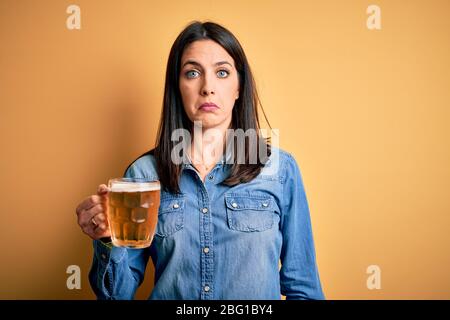 Young woman with blue eyes drinking jar of beer standing over isolated yellow background depressed and worry for distress, crying angry and afraid. Sa Stock Photo