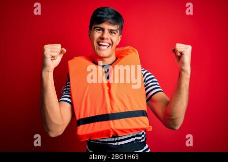 Young handsome man wearing orange safety life jacket over isolated red background celebrating surprised and amazed for success with arms raised and op Stock Photo