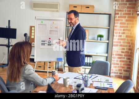 Two middle age business workers smiling happy and confidentt working together with smile on face. Man standing doing work presentation using board and Stock Photo