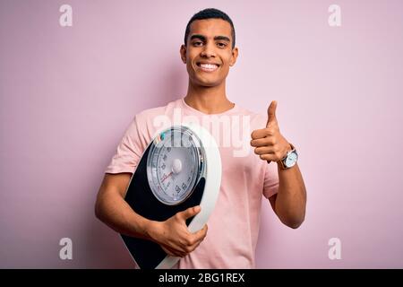 Young handsome african american slim sportsman using weight machine over pink background happy with big smile doing ok sign, thumb up with fingers, ex Stock Photo