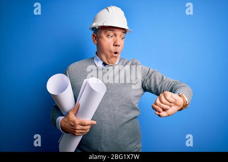 Middle age handsome grey-haired architect man wearing safety helmet holding blueprints Looking at the watch time worried, afraid of getting late Stock Photo