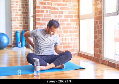 Middle age handsome sportman sitting on mat doing stretching yoga exercise at gym Suffering of backache, touching back with hand, muscular pain Stock Photo