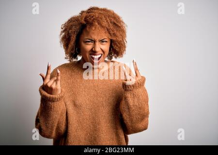 Young beautiful African American afro woman with curly hair wearing casual sweater shouting with crazy expression doing rock symbol with hands up. Mus Stock Photo