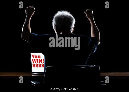 Back view of a happy man with gray hair  working at night, raising up his hands in excitement, looking at his computer screen with You Win! $$$ text w Stock Photo