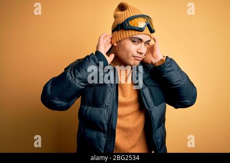 Young brazilian skier man wearing snow sportswear and ski goggles over yellow background Trying to hear both hands on ear gesture, curious for gossip. Stock Photo