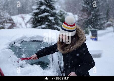 Young attractive brunette woman clearing snow off a car with a ice scraper in the winter. Stock Photo